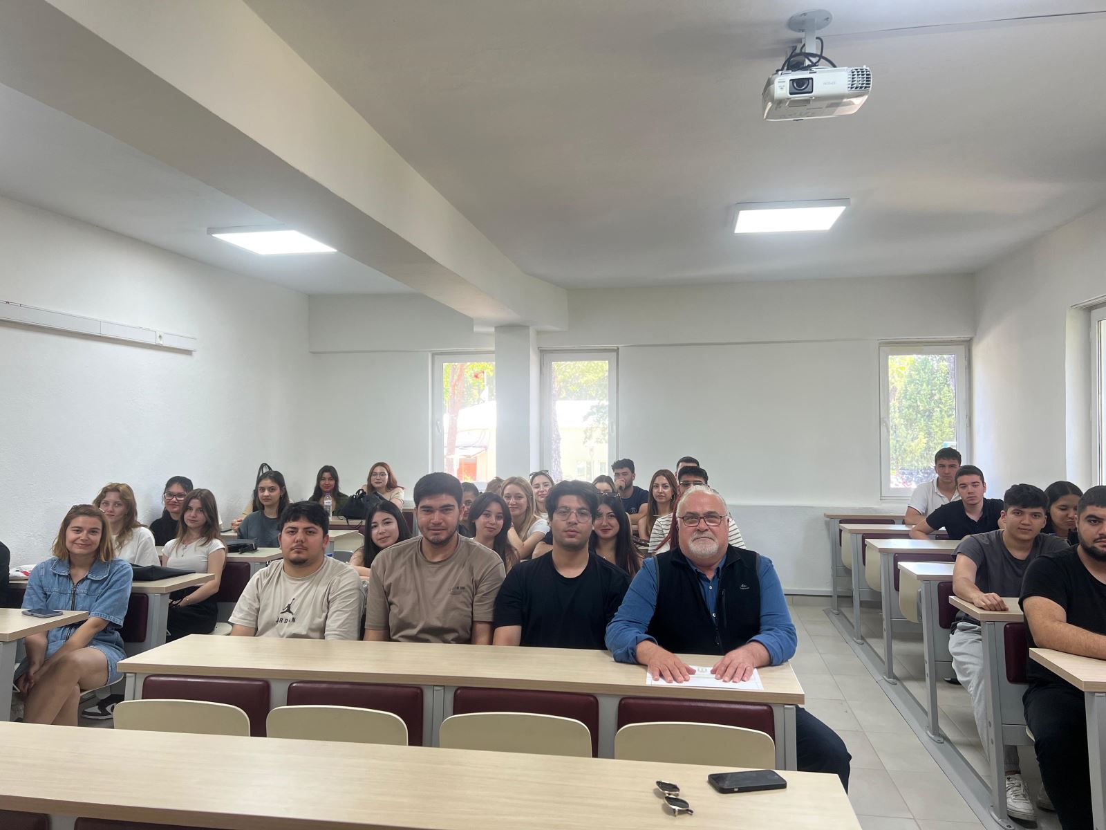 Aydınlık bir sınıf ortamında çekilen bu fotoğrafta, orta yaşlı bir erkek (konferans konuşmacısı) ön sırada ortada oturmakta. Sağında ve solunda öğrenciler yer almakta. Arka sıralarda da yaklaşık 30’a yakın öğrenci sırayla oturmuş, objektife doğru bakıyorlar. Öğrencilerin çoğu gülümsüyor. Sınıfın tavanında bir projeksiyon cihazı, duvarlarında ise büyük pencereler yer alıyor. Fotoğraf, derslikteki samimi ve dikkatli atmosferi yansıtıyor.